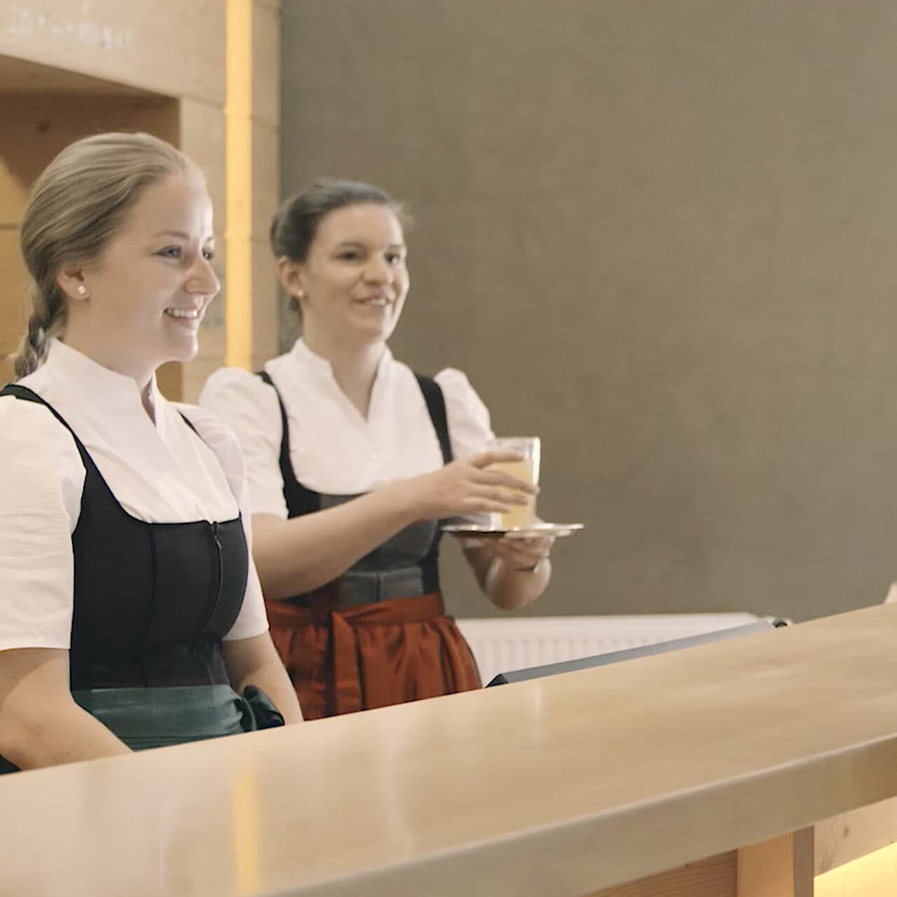 Zwei Frauen in traditioneller Kleidung stehen hinter einem Empfangstresen vor einem galerieähnlichen, neutralen Hintergrund. Die eine lächelt herzlich, die andere hält anmutig ein Tablett mit Gläsern.