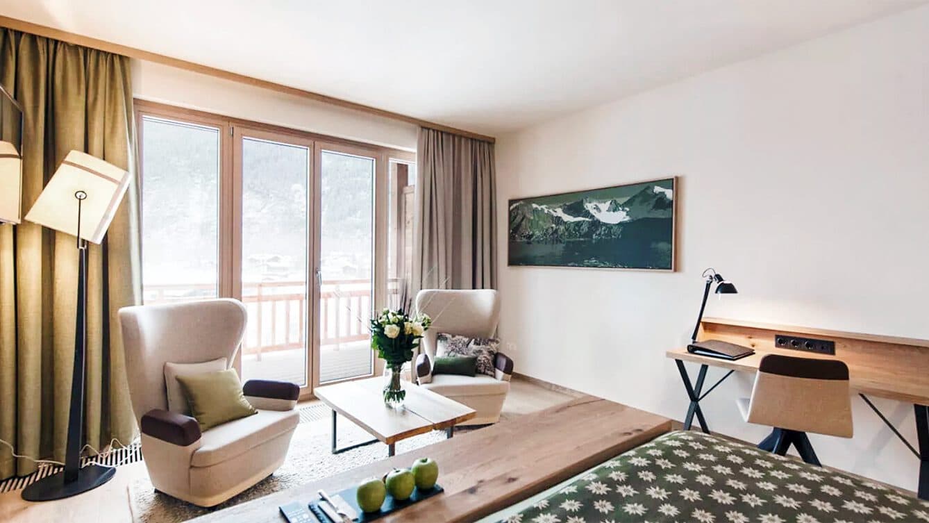 Modernes Hotelzimmer mit zwei Sesseln, einem Tisch, einem Schreibtisch und einem großen Fenster mit Bergblick. Ein Bett mit gemusterter Tagesdecke harmoniert mit dem gerahmten Kunstwerk an der Wand und verleiht dem Raum eine galerieartige Atmosphäre.