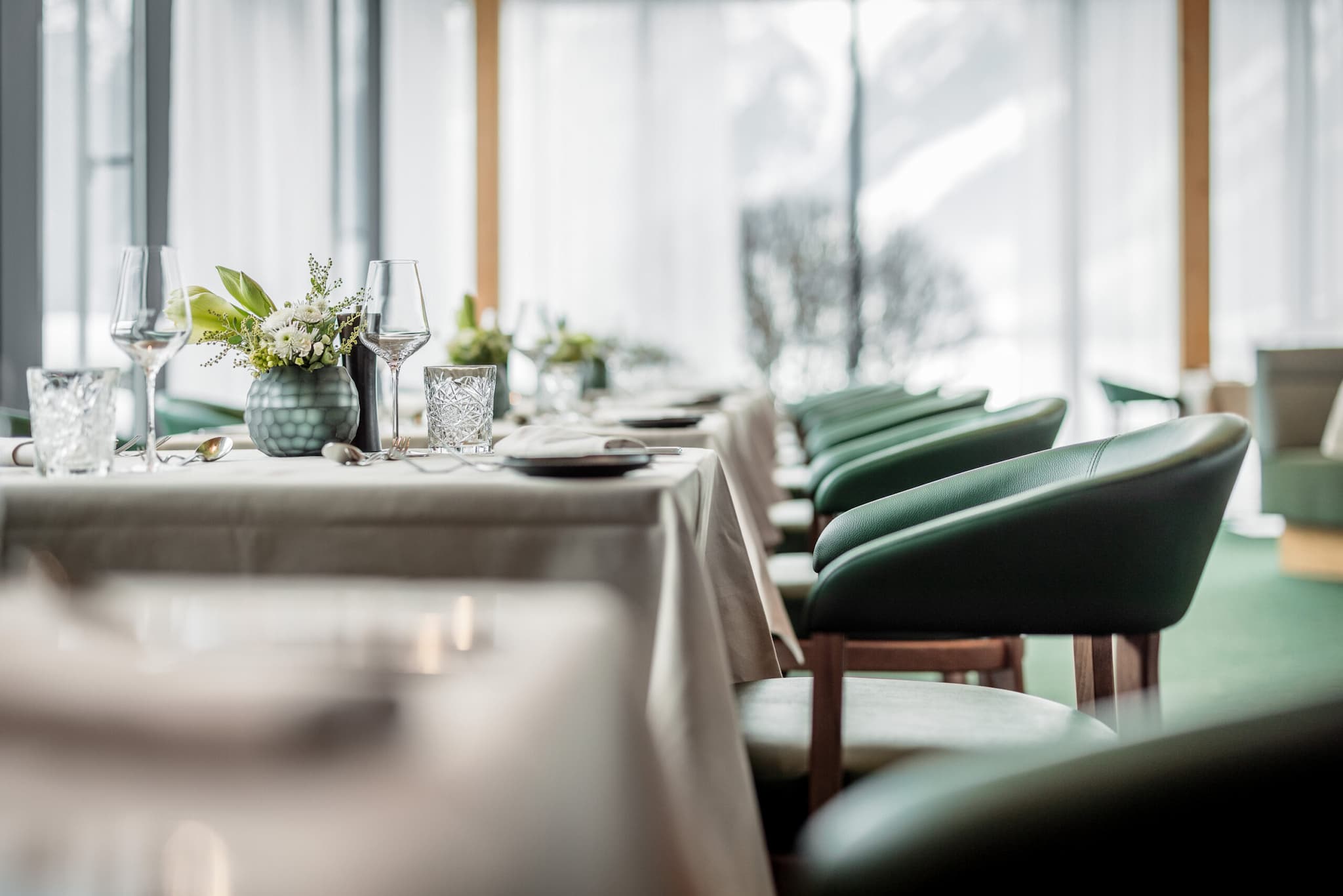 Elegant gedeckte Restauranttische mit weißen Tischdecken, Weingläsern und Blumenschmuck, umgeben von grünen Stühlen. Natürliches Licht fällt durch große Fenster.