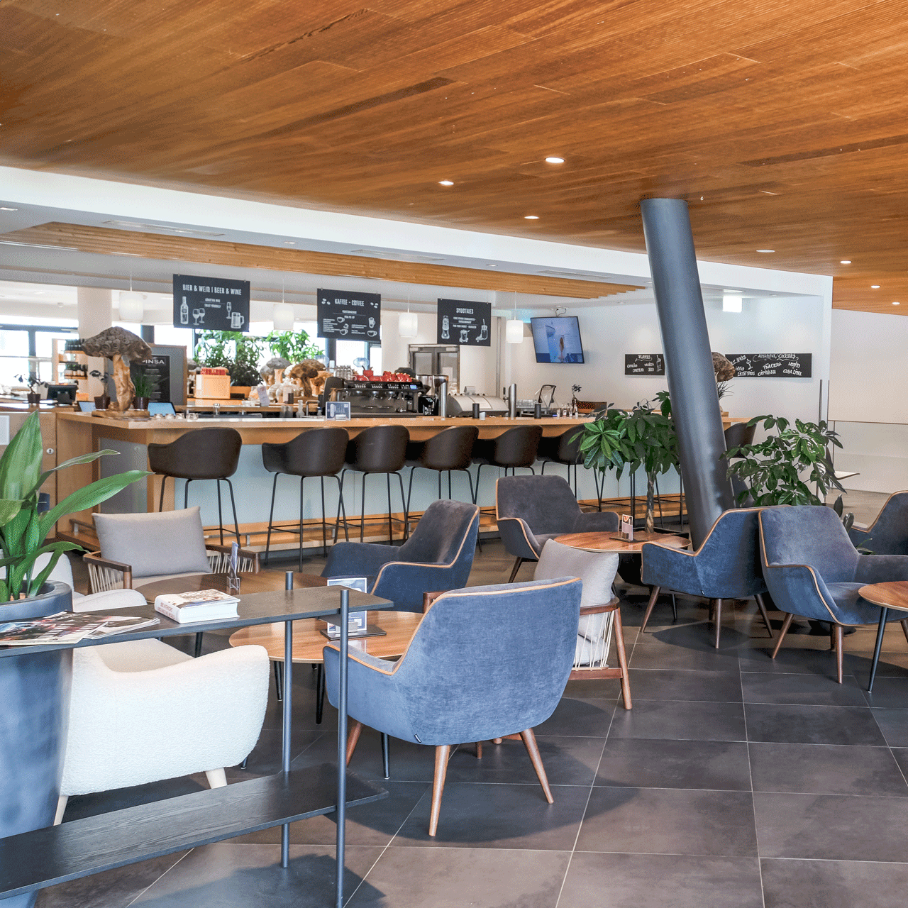 Modern cafe interior with wooden ceiling and stone tile floor. Seating includes gray and beige chairs, round tables, and a bar with stools. Green plants add to the decor.