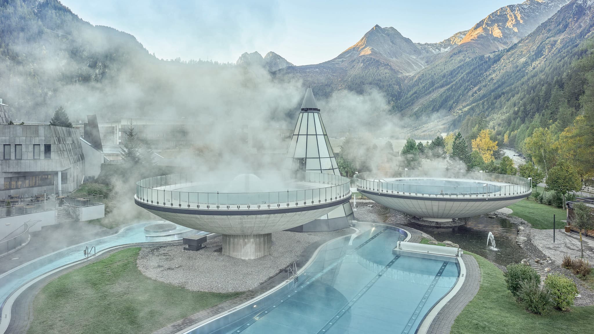 Futuristic buildings with steam rising, nestled in a mountainous landscape, feature two large, round structures and a long pool. Snow-capped peaks and greenery are visible in the background.