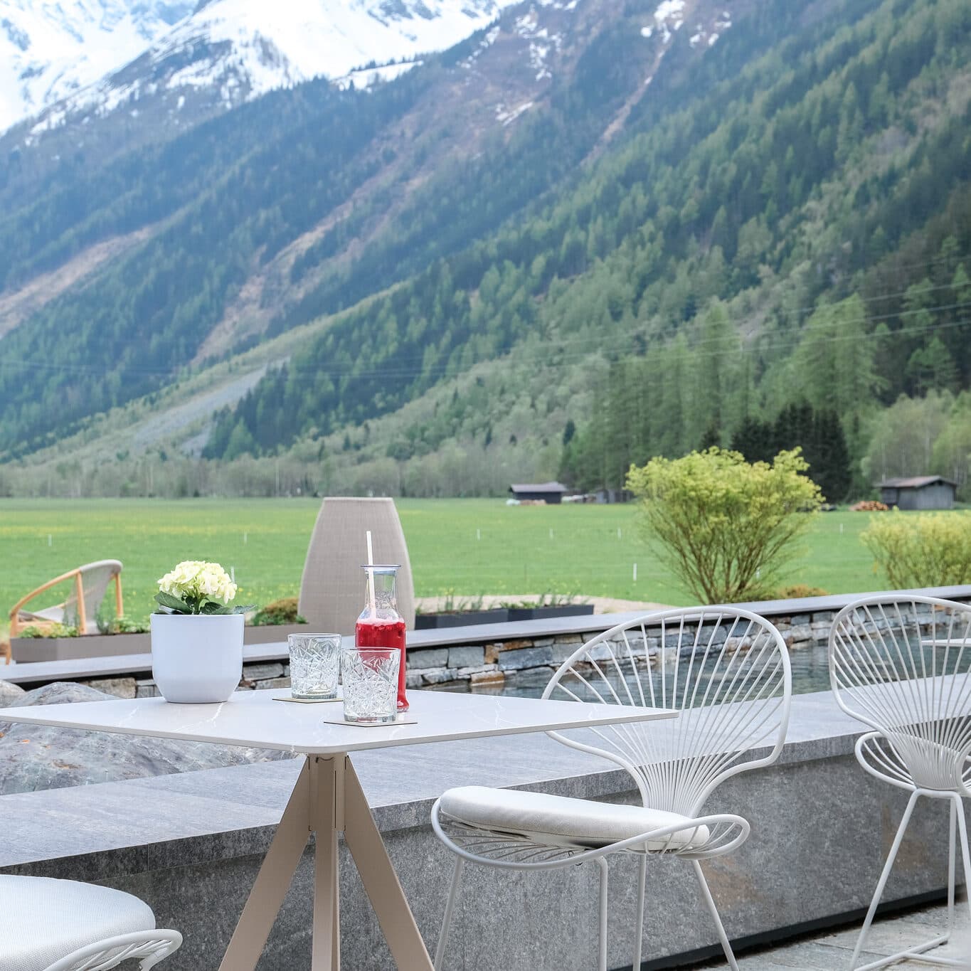 Il patio esterno del ristorante è arredato con eleganti sedie in filo metallico e un tavolo decorato con bevande, fiori e bicchieri, e offre una vista mozzafiato sui campi verdi e sulle montagne boscose con le cime innevate in lontananza.