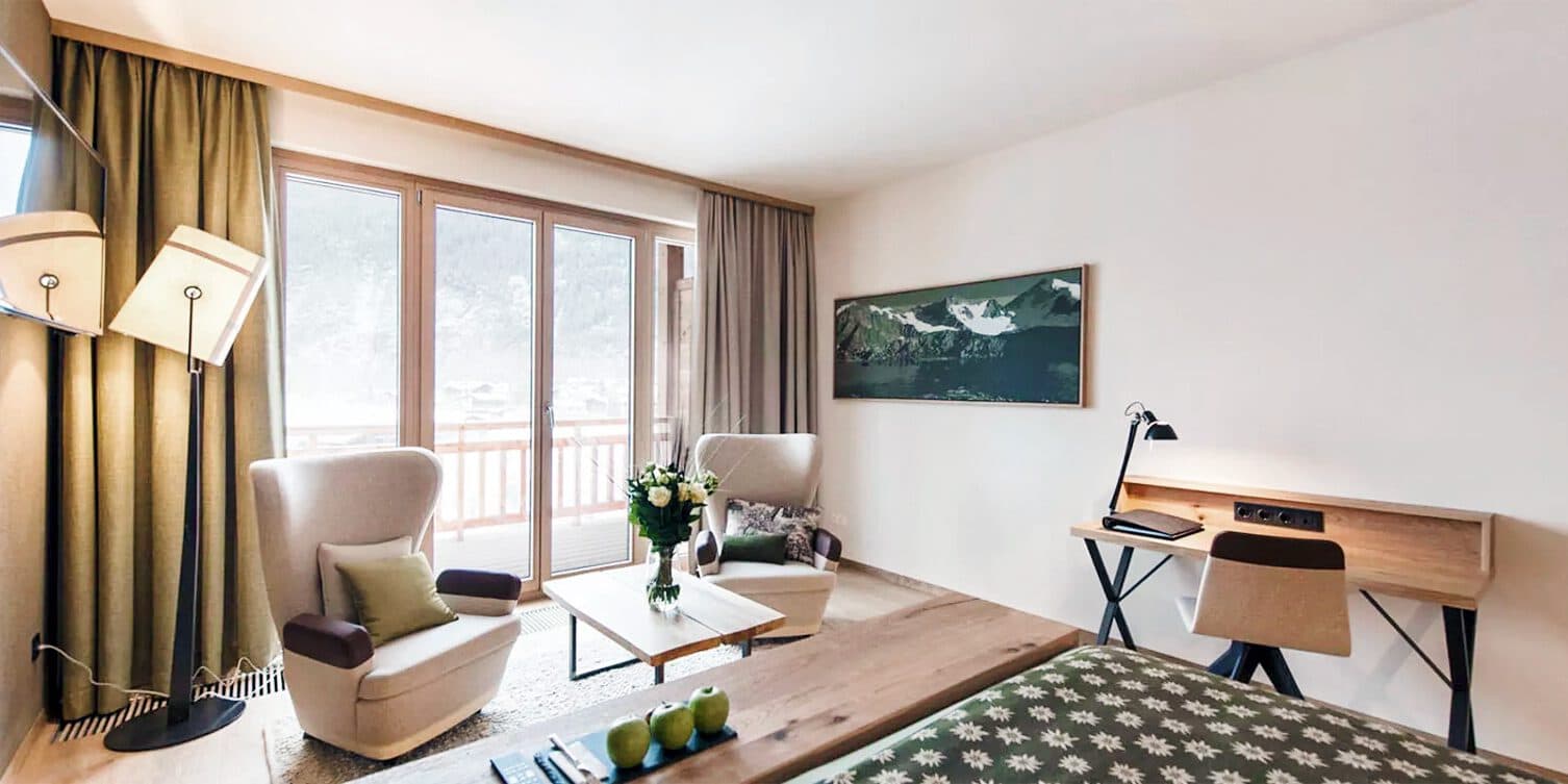 Modern hotel room with two chairs and a table near a large window. A desk with a lamp is on the right, and a bed is partially visible. Walls are decorated with a mountain landscape photo.