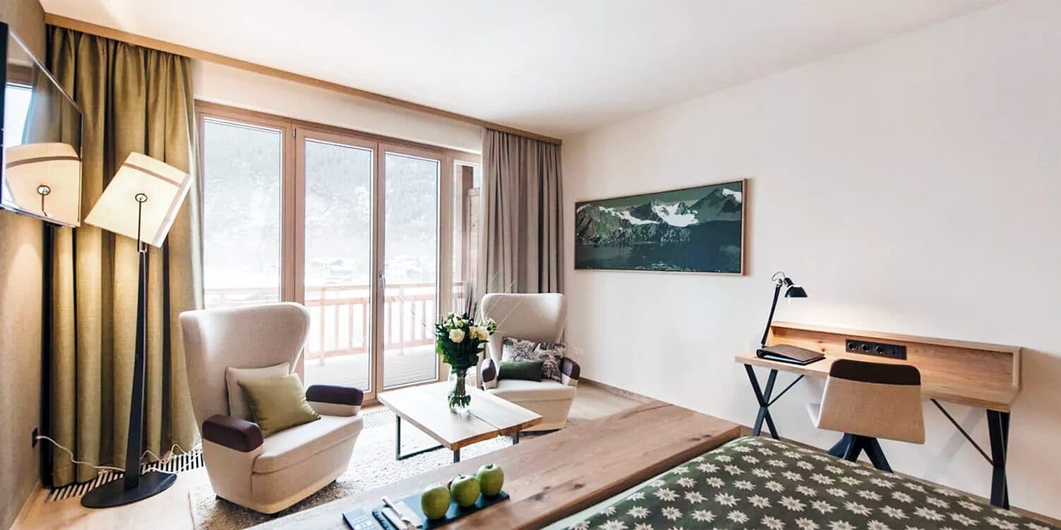Moderna camera d'albergo con due poltrone, un tavolo in legno e un letto. Una scrivania con lampada è posizionata vicino alla parete. Un'ampia finestra con tende offre una vista sull'esterno. La parete decorata con un paesaggio montano.
