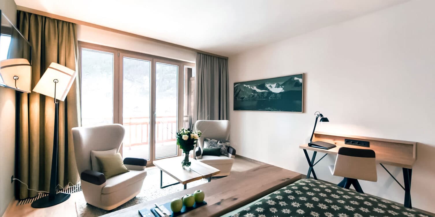 Chambre d'hôtel moderne avec deux fauteuils et une table, un bureau avec une chaise, un lit, une télévision murale et une fenêtre avec vue sur le balcon.
