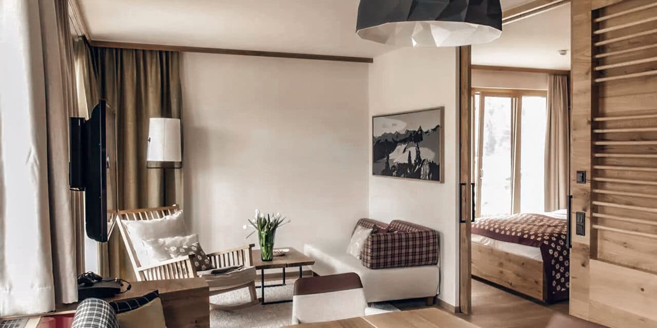 Un'accogliente zona giorno con un piccolo divano e una poltrona, un tavolino da caffè in legno e una camera da letto con letto visibile attraverso una porta scorrevole. L'arredamento include un dipinto murale e un vaso per piante.