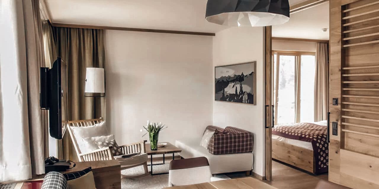 Un salon chaleureux avec un fauteuil moderne, un petit canapé et une table basse fleurie, adjacent à une chambre visible par une porte coulissante. Des tons neutres et une lumière naturelle imprègnent l'espace.