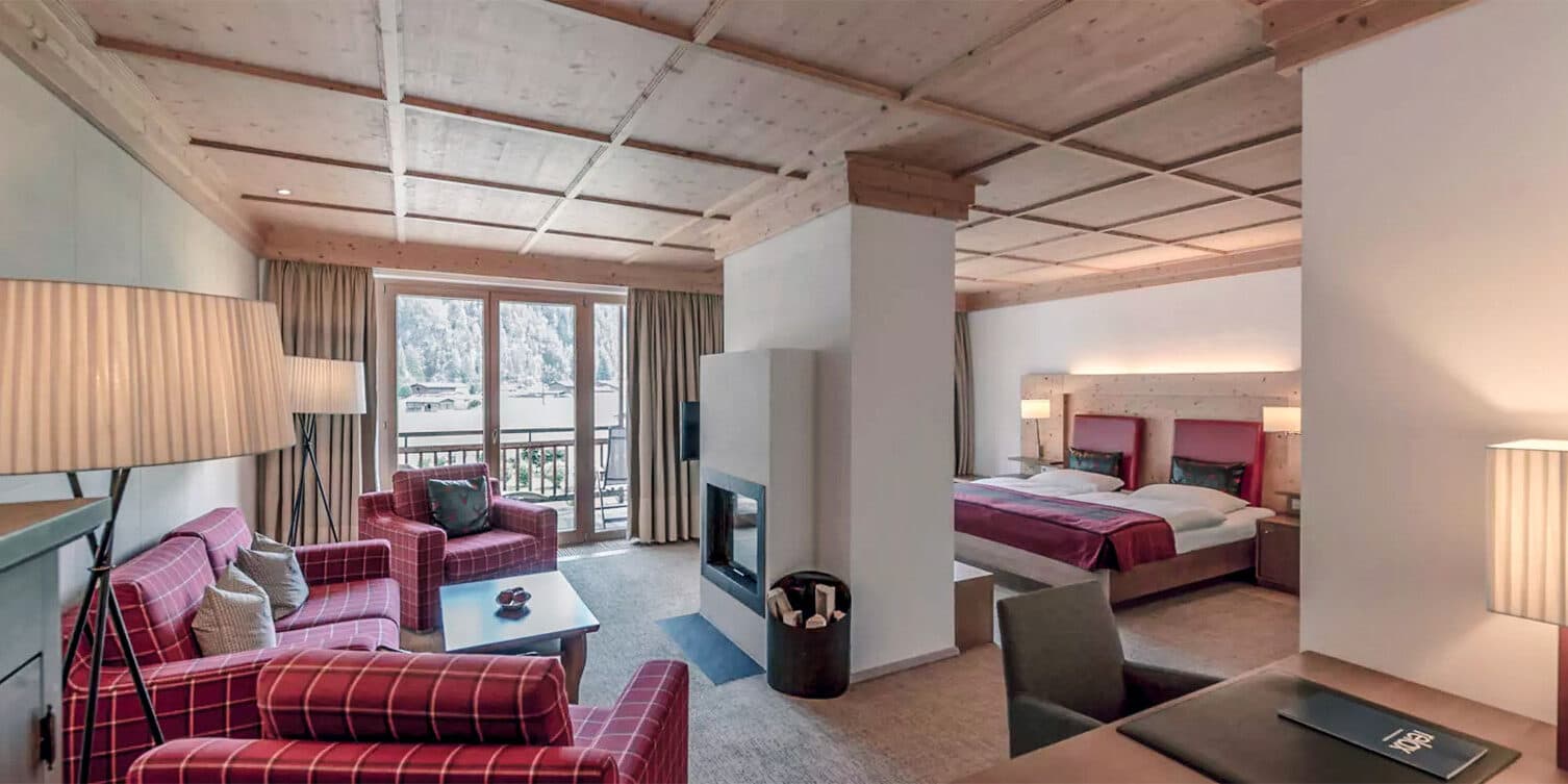 Suite d'hôtel avec deux lits, un coin salon confortable avec des chaises à carreaux rouges, une cheminée, un bureau et de grandes fenêtres avec vue sur la montagne.