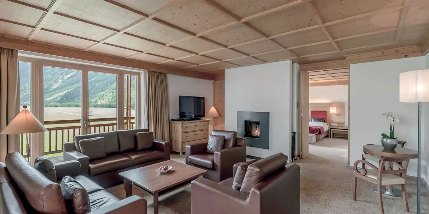 Suite d'hôtel moderne avec canapés en cuir marron, télévision et cheminée. De grandes fenêtres offrent une vue sur les montagnes. Chambre avec décoration en bois visible par une porte ouverte.