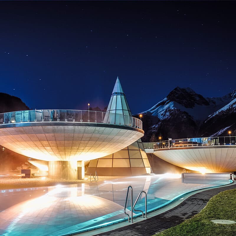 Ein moderner, beleuchteter Day-Spa-Komplex mit geometrischen Kuppelstrukturen und einem Außenpool bei Nacht vor der Kulisse schneebedeckter Berge.