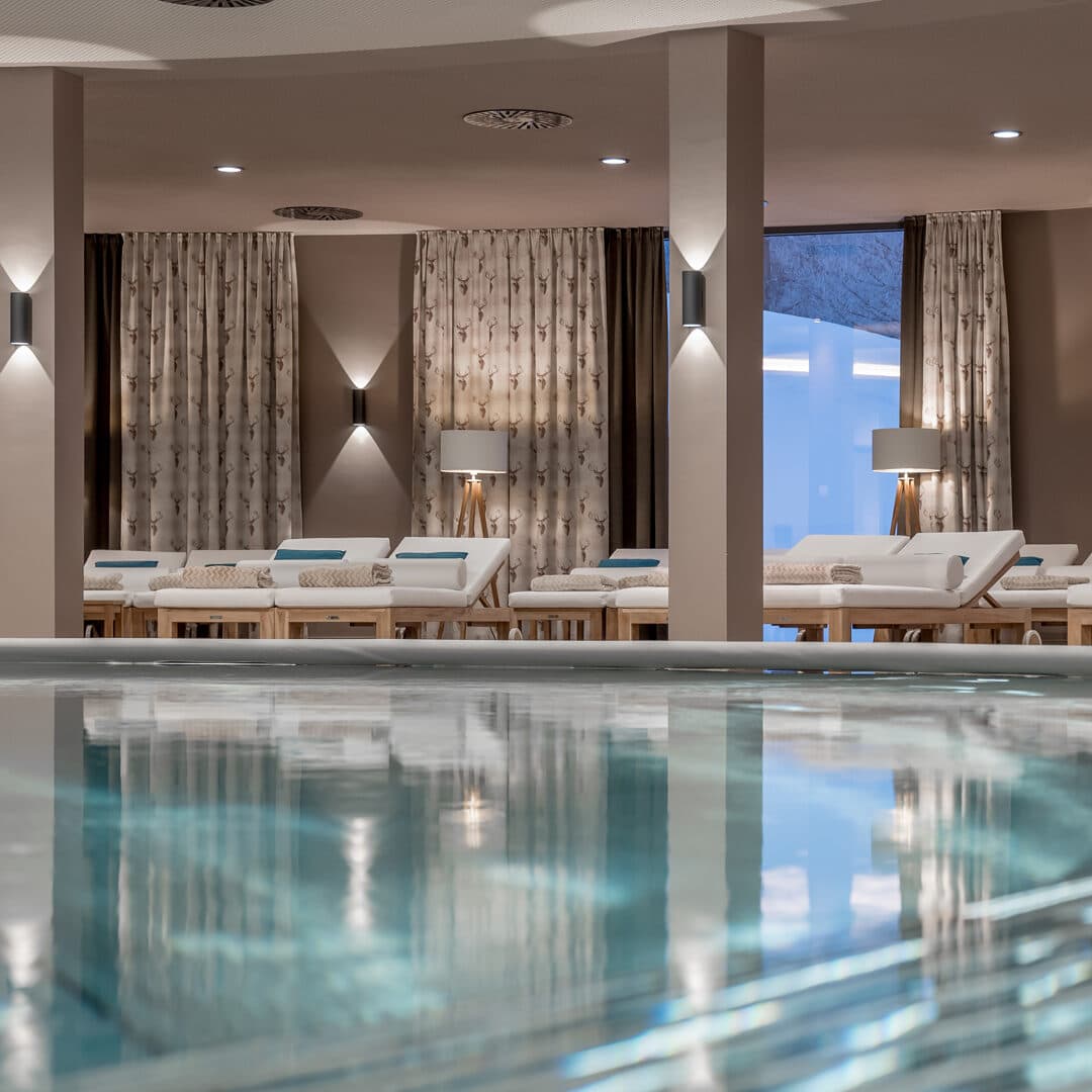 Der Innenbereich des Day Spas besticht durch einen einladenden Pool im Vordergrund. Mehrere Liegestühle mit Kissen säumen die Rückwand, begleitet von Stehlampen und gemusterten Vorhängen, die eine ruhige Atmosphäre schaffen.