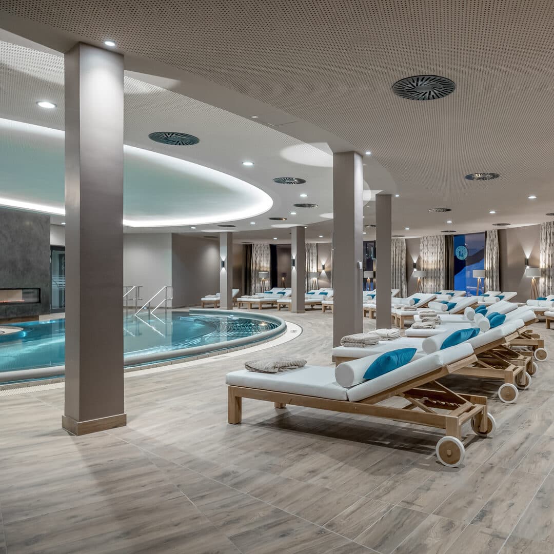 Betreten Sie unser ruhiges Day Spa mit einem runden Innenpool, umgeben von Liegestühlen mit weißen und blauen Kissen. Der Raum besticht durch sein modernes Design mit Holzboden und sanfter Beleuchtung – eine Oase der Entspannung.