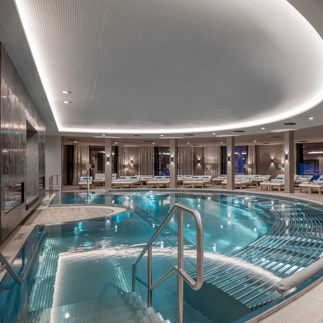 Innenpool mit modernem Design, Metallgeländern, einem zentralen runden Whirlpool, Liegestühlen am Rand und sanfter Umgebungsbeleuchtung, die eine ruhige Day-Spa-Atmosphäre schaffen.