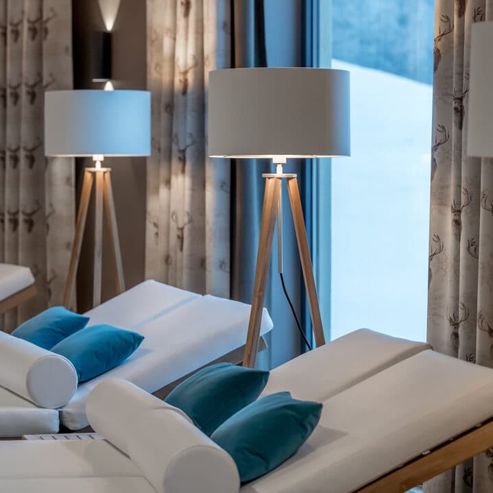 Der Loungebereich des Day Spas verfügt über zwei weiße Chaiselongues, jeweils mit türkisfarbenen Kissen ausgestattet. Neben den Liegen stehen Holzlampen mit Dreibeinen, während ein Fenster den Blick auf eine ruhige, verschneite Außenlandschaft freigibt.