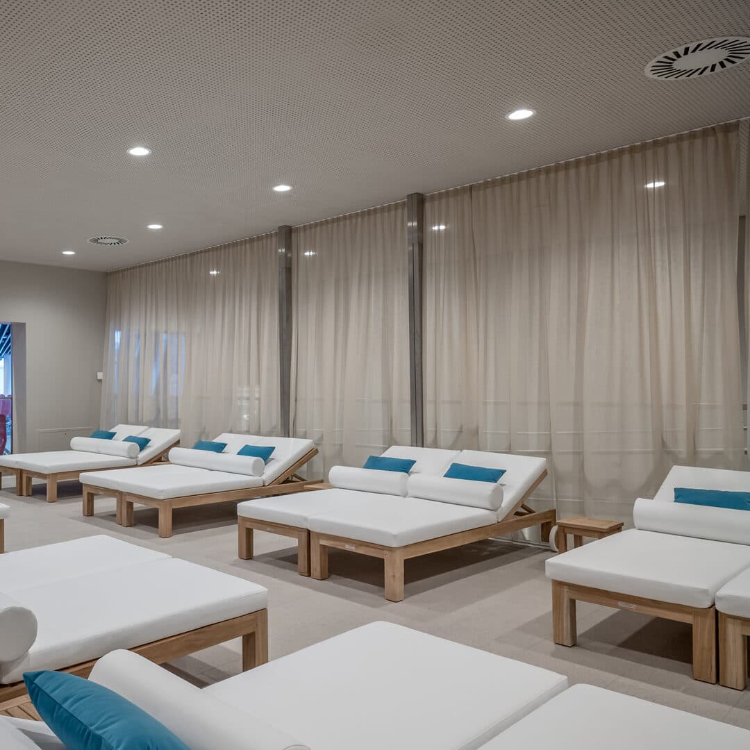 Ein moderner Entspannungsraum eines Day Spas mit mehreren weißen Liegestühlen, jeder mit einem blauen Kissen ausgestattet, unter einer sanft beleuchteten Decke und im Hintergrund von beigen Vorhängen eingerahmt.