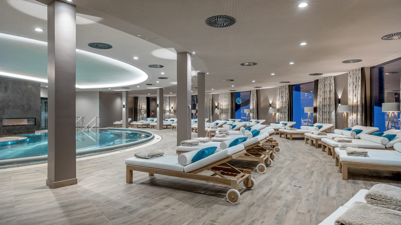 Luxuriöses Spa-Interieur mit rundem Innenpool, umgeben von Holzliegen mit weißen Kissen und gerollten Handtüchern. Sanfte Beleuchtung und modernes Design schaffen eine entspannende Atmosphäre.
