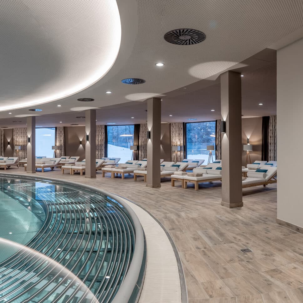 Moderner Hallenbadbereich mit Holzboden, mehreren Liegestühlen und großen Fenstern mit Blick ins Freie.