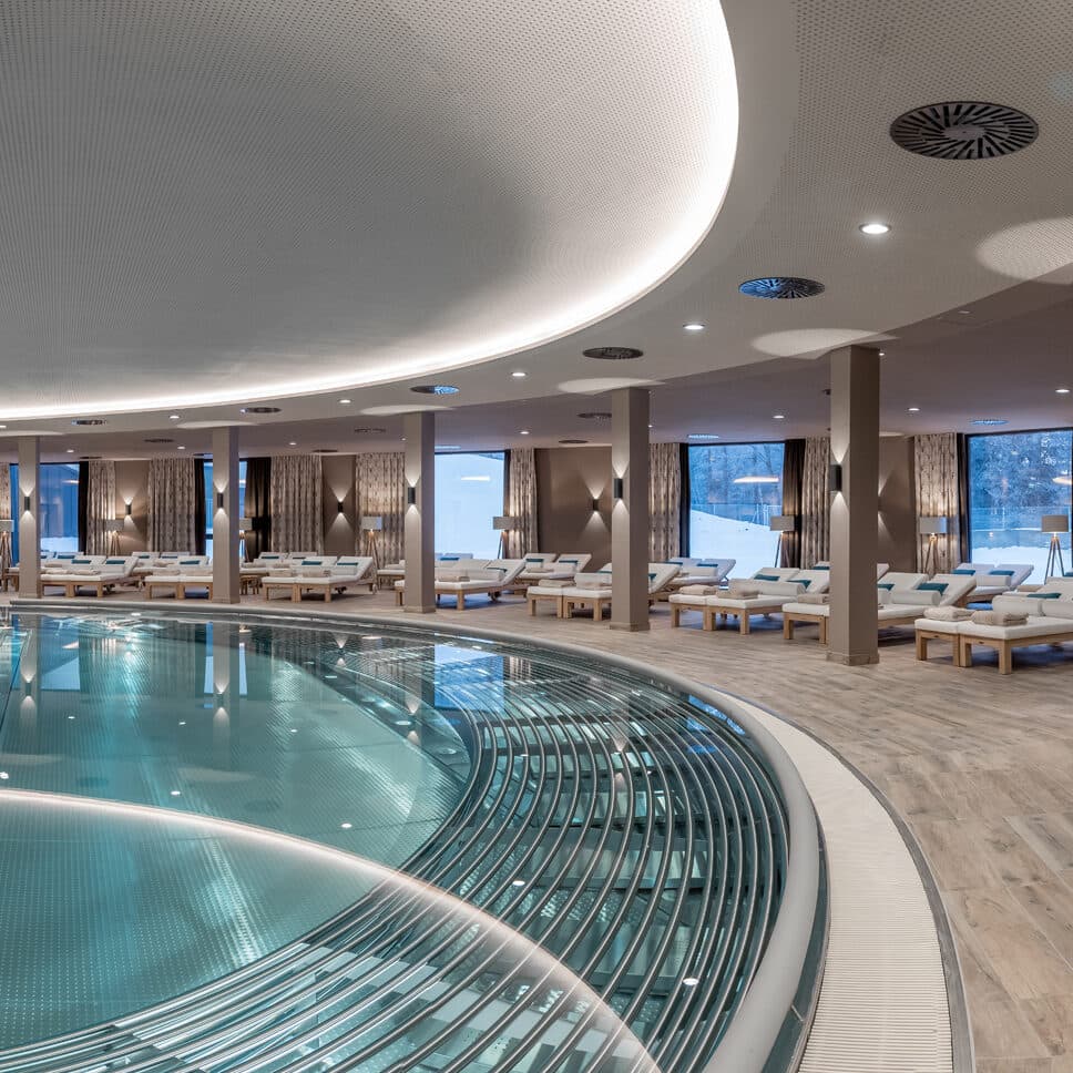Indoor spa with a large circular pool, lounge chairs, and floor-to-ceiling windows showing an outdoor view. The space features modern lighting and wooden flooring.