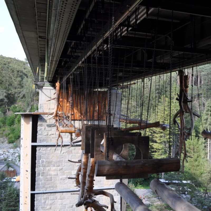 An der Unterseite einer Brücke hängen mit Ketten befestigte Holzschaukeln und -konstruktionen, im Hintergrund sind Bäume und eine Steinsäule zu sehen.
