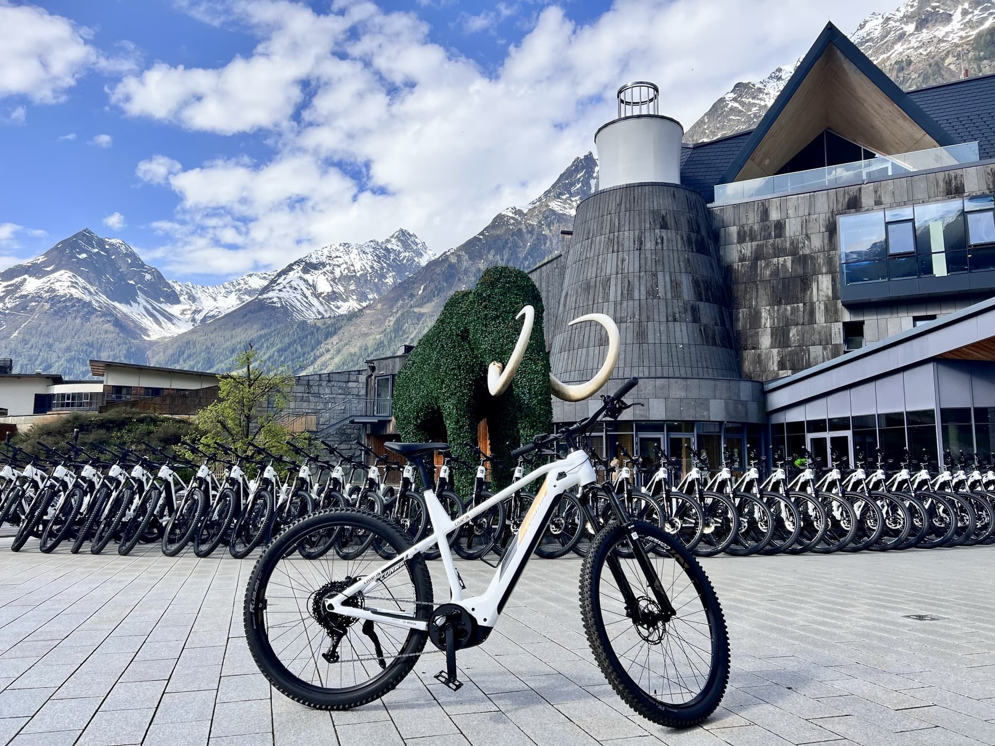 Ein weißes Mountainbike steht auf einer gepflasterten Fläche vor einem modernen Gebäude, im Hintergrund sind schneebedeckte Berge und ein Fahrradständer zu sehen.