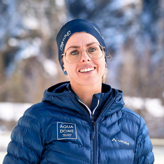 Eine Person mit Brille, einer blauen Winterjacke mit Sponsorenlogos und einer blauen Mütze steht im Freien in einer verschneiten Landschaft.