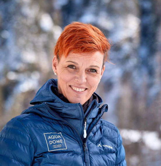 Eine Person mit kurzen orangefarbenen Haaren in einer blauen Jacke steht im Freien vor einem verschneiten, unscharfen Hintergrund. Die Jacke trägt die Logos von AQUA DOME und VAUDE.