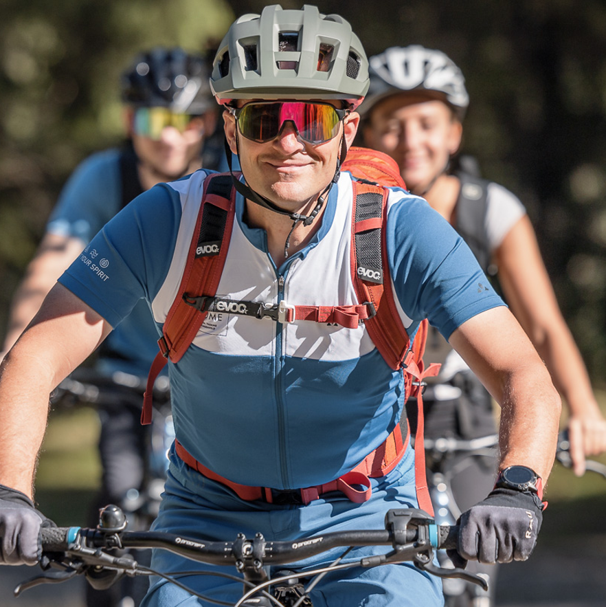 Trois cyclistes portant des casques et des lunettes de soleil font du vélo à l'extérieur par une journée ensoleillée, la personne à l'avant souriant à l'appareil photo.