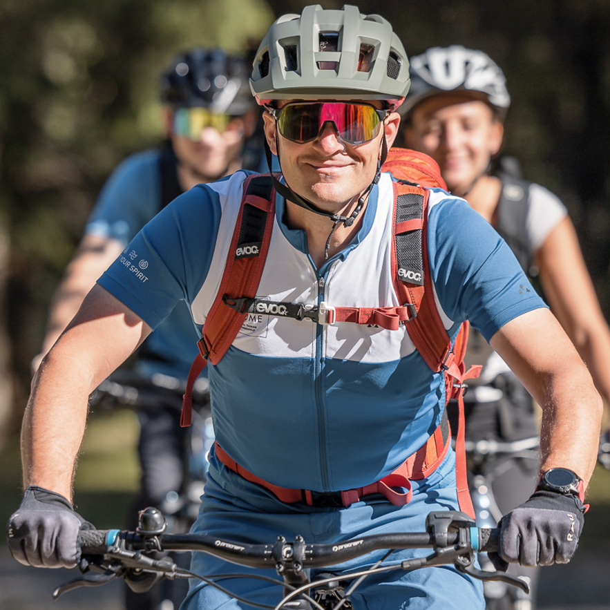 Drei Radfahrer mit Helmen, Sonnenbrillen und Radsportbekleidung fahren im Freien in einer Reihe, wobei der führende Radfahrer klar zu sehen ist und die anderen im Hintergrund leicht verschwommen sind.