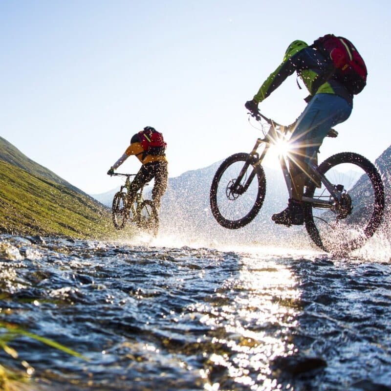 Zwei Radfahrer fahren an einem sonnigen Tag mit Mountainbikes durch einen seichten Bach, wobei das Wasser spritzt und das Sonnenlicht durch ihre Räder scheint.