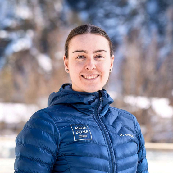 Eine Person in einer blauen Winterjacke steht im Freien vor einem verschwommenen Schnee- und Berghintergrund und lächelt in die Kamera.