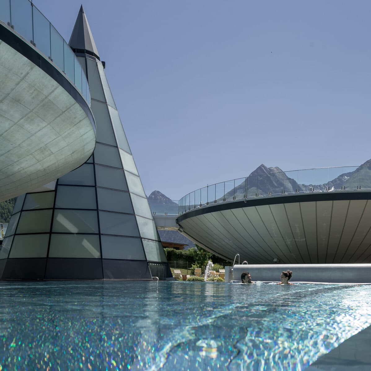 Zwei Personen schwimmen in einem modernen Freibad mit pyramiden- und schüsselförmigen Strukturen aus Glas und Beton in der Nähe, im Hintergrund sind bei klarem Himmel die Berge zu sehen.