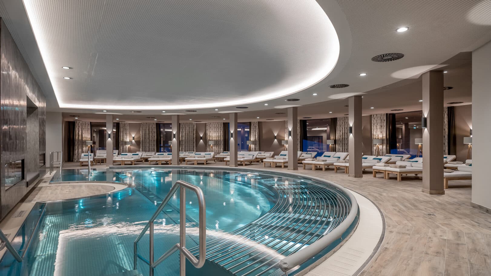 Indoor spa with a curved pool, lounge chairs along the sides, modern lighting, and neutral-toned decor.