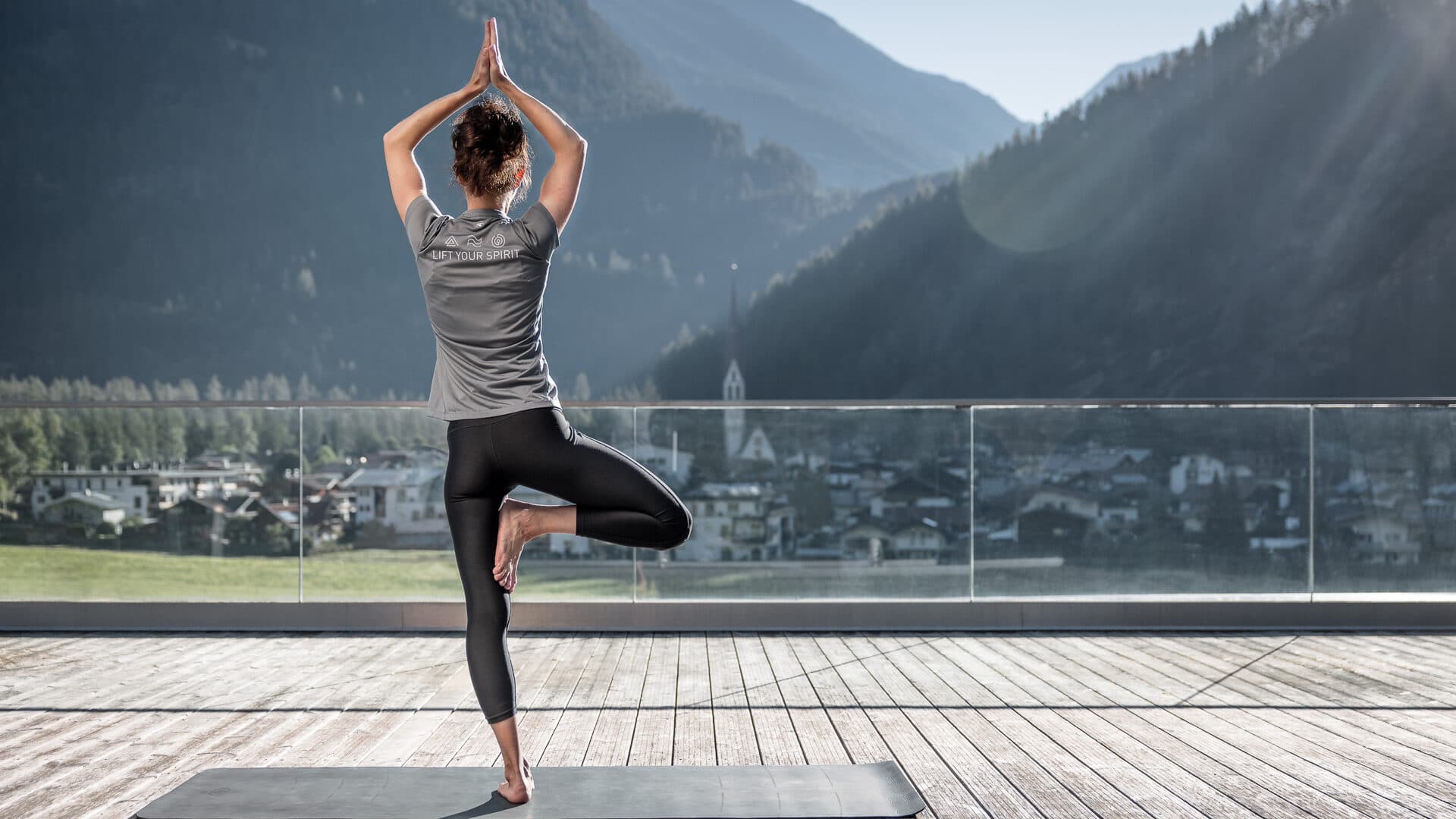 Eine Person übt auf einer Matte im Freien Yoga in der Baumstellung, während im Hintergrund bei klarem Himmel Berge und eine Stadt zu sehen sind.