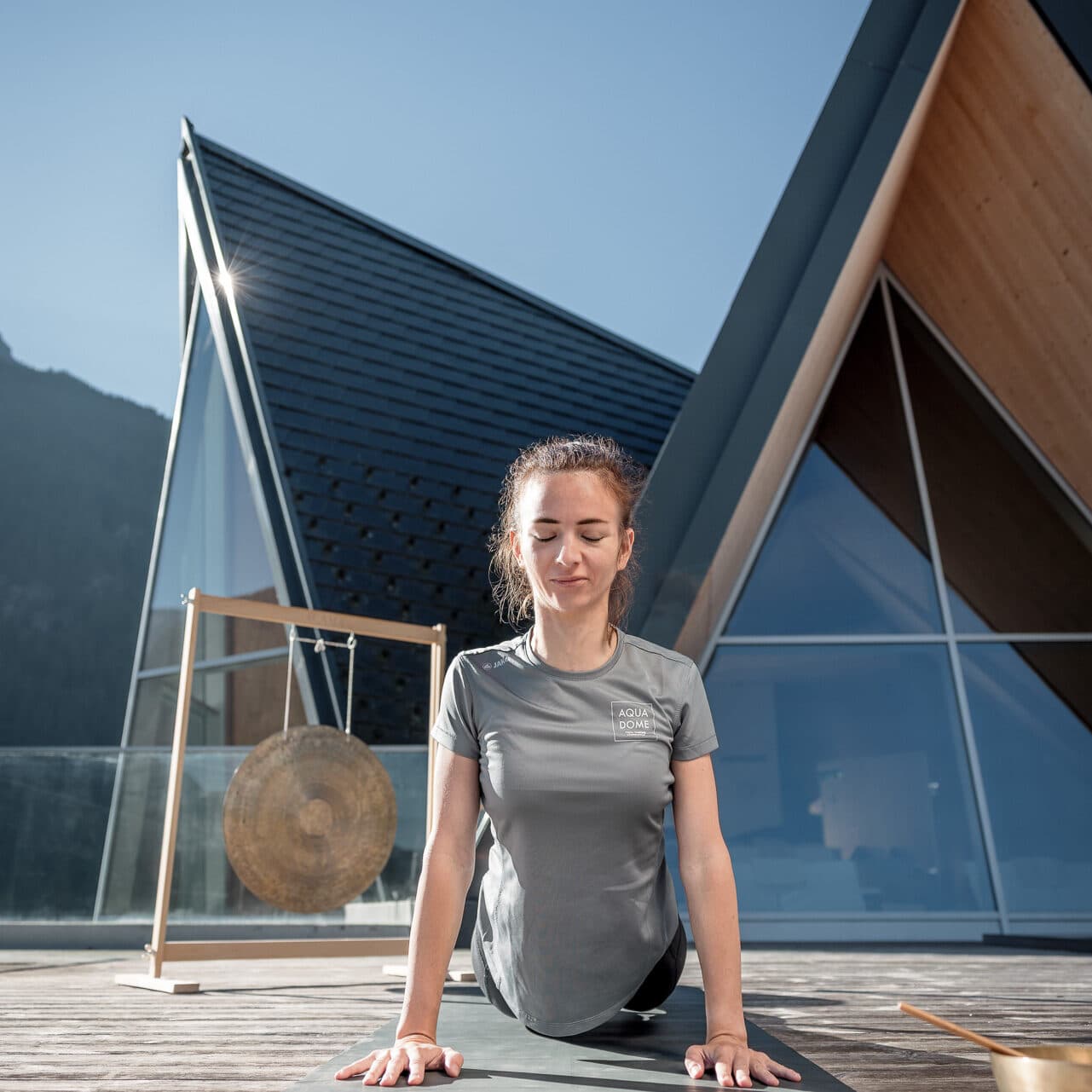 Eine Frau praktiziert Yoga im Freien auf einer Matte und nimmt vor einem modernen, kantigen Gebäude mit großen Fenstern eine nach oben gerichtete Haltung ein.