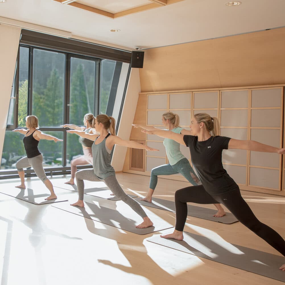 Fünf Personen, die in einem hellen Studio Yoga praktizieren, stehen alle auf Matten und nehmen eine Kriegerpose mit ausgestreckten Armen ein.