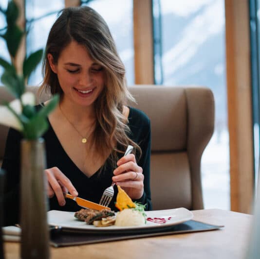 Eine Frau sitzt an einem Tisch in einem Restaurant und schneidet lächelnd ihr Essen mit Messer und Gabel. Vor ihr steht ein Teller mit verschiedenen Speisen, und ein Fenster zeigt eine verschneite Landschaft draußen.