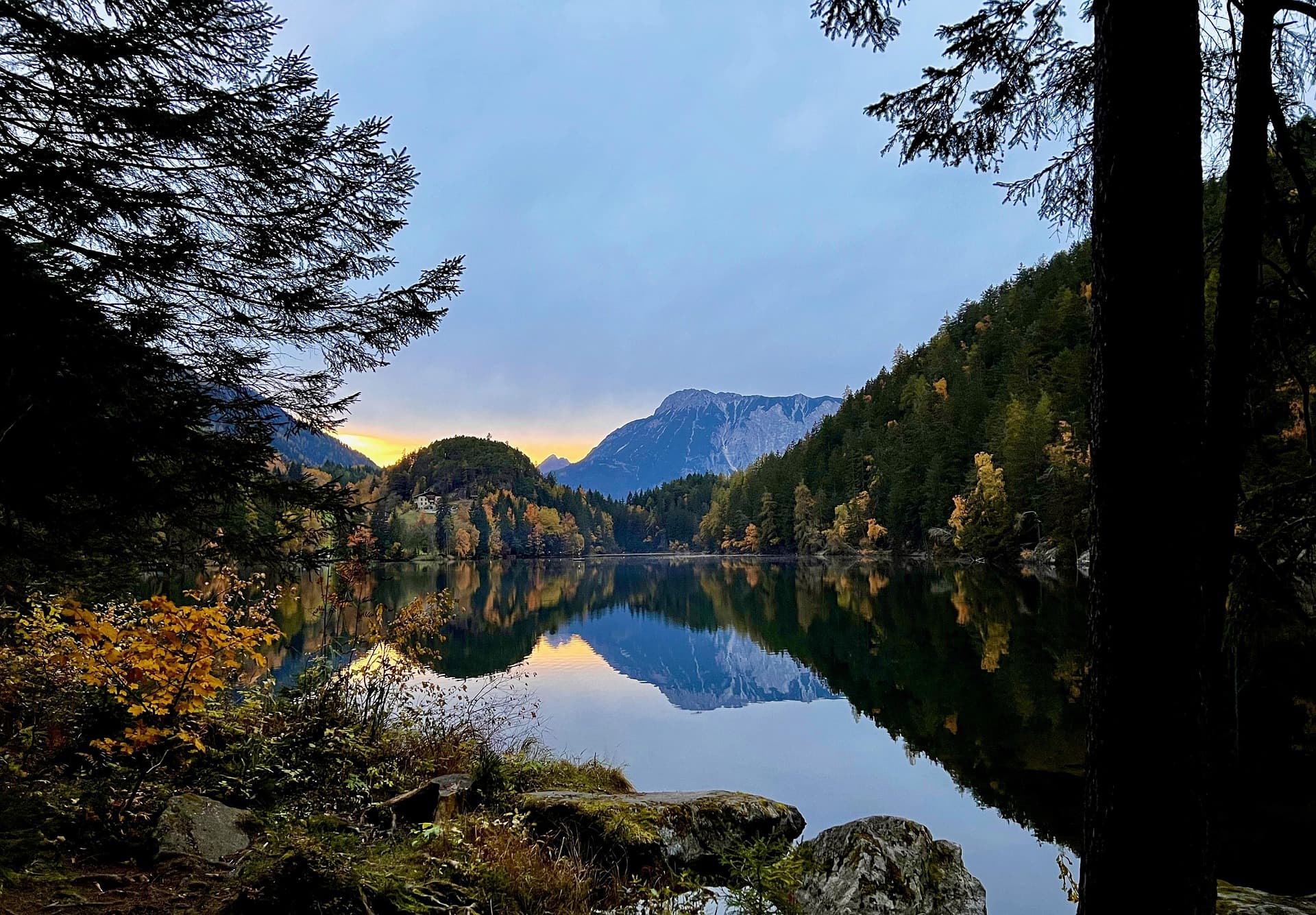 Ein ruhiger See, umgeben von herbstlichen Bäumen und Bergen, in dem sich die Landschaft und der Himmel spiegeln.