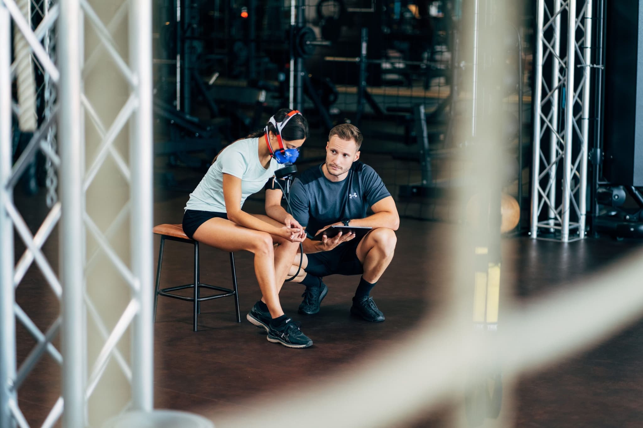 Eine Person, die Sportkleidung und eine Atemmaske trägt, sitzt auf einer Bank, während eine andere Person neben ihr kniet. Beide schauen auf ein Tablet, während sie die Ergebnisse der uVida Atem-Stoffwechselanalyse in einem Fitnessstudio überprüfen.