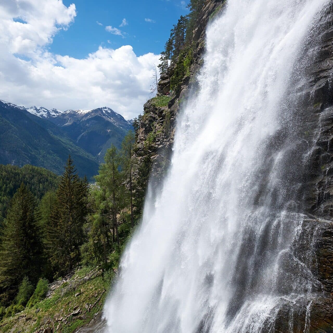 Großer Wasserfall, der eine von Kiefern umgebene Felswand hinabstürzt, mit Bergen und einem teilweise bewölkten Himmel im Hintergrund.