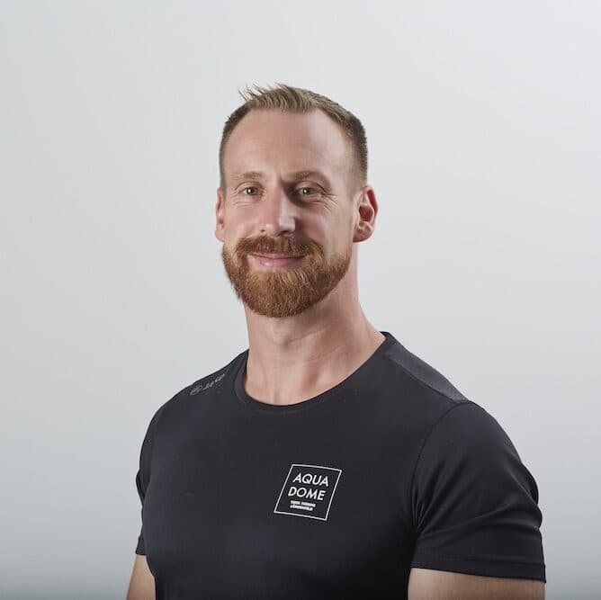 Ein Mann mit gepflegtem Bart und kurzen Haaren, der ein schwarzes Aqua-Dome-T-Shirt trägt, steht vor einem schlichten, hellen Hintergrund und schaut in die Kamera.