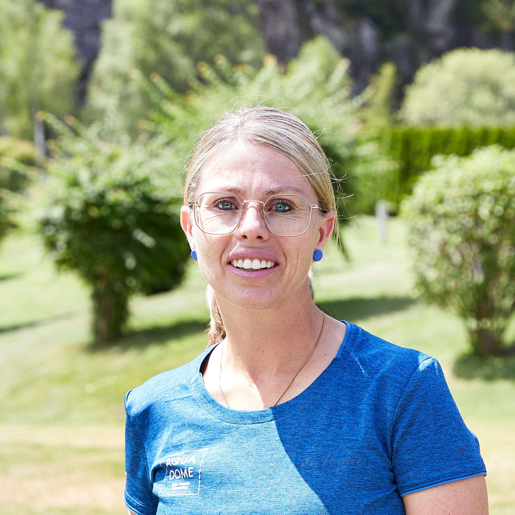 Eine Frau mit blondem Haar, Brille und blauem Hemd steht draußen in einem sonnigen, grünen Park.