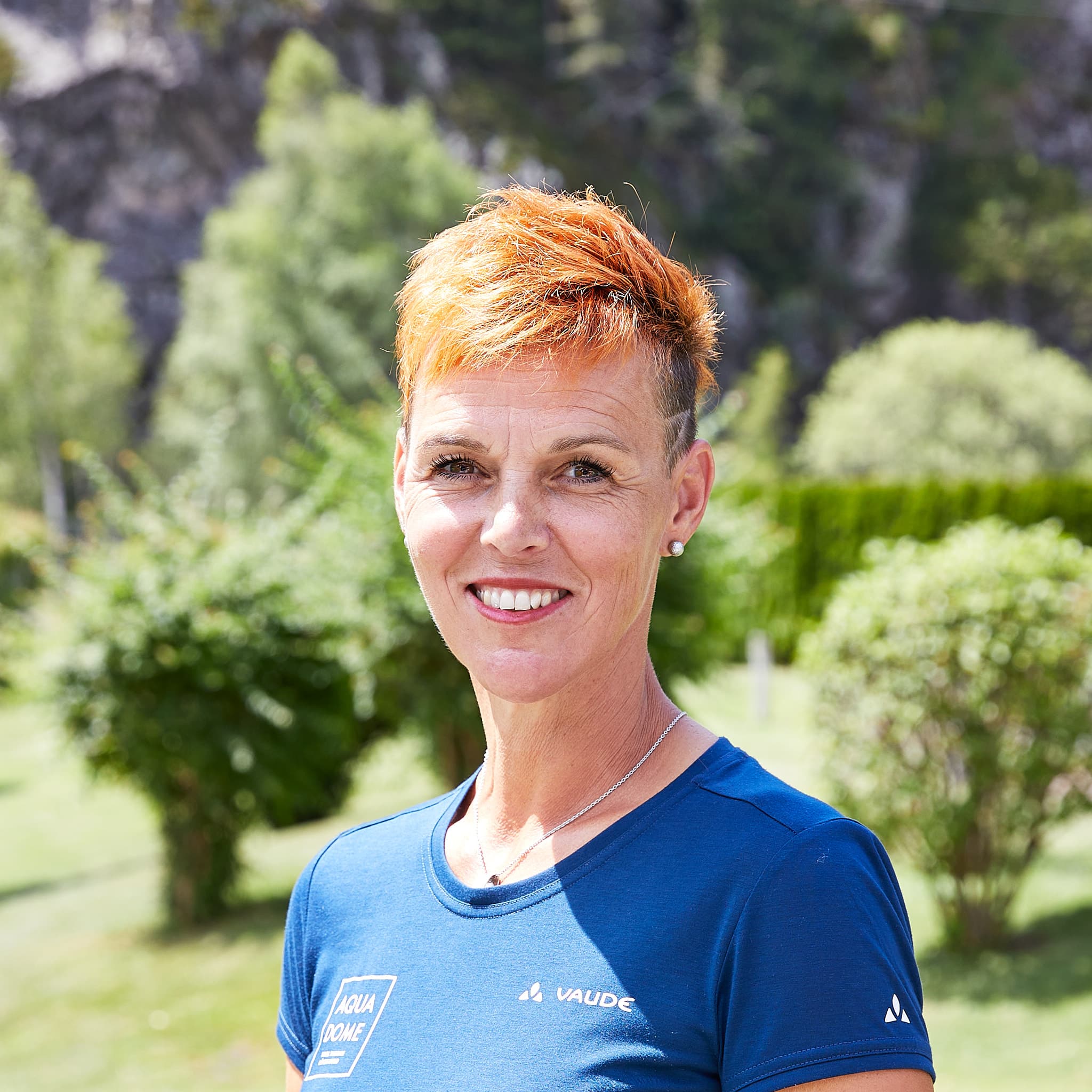 Eine Frau mit kurzen orangefarbenen Haaren und einem blauen T-Shirt lächelt im Freien mit Bäumen und Grünpflanzen im Hintergrund.