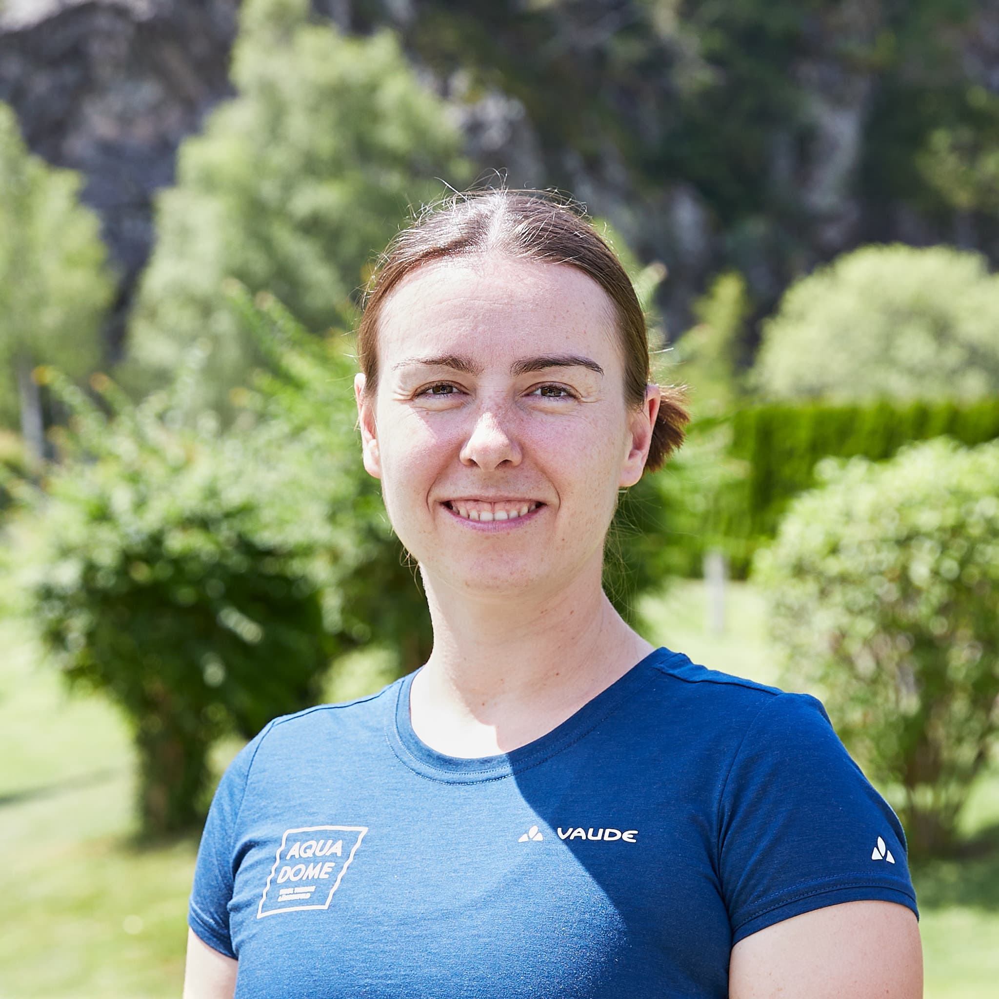 Eine Frau mit zurückgebundenen braunen Haaren und blauem T-Shirt steht im Freien, mit Grün und Bäumen im Hintergrund.
