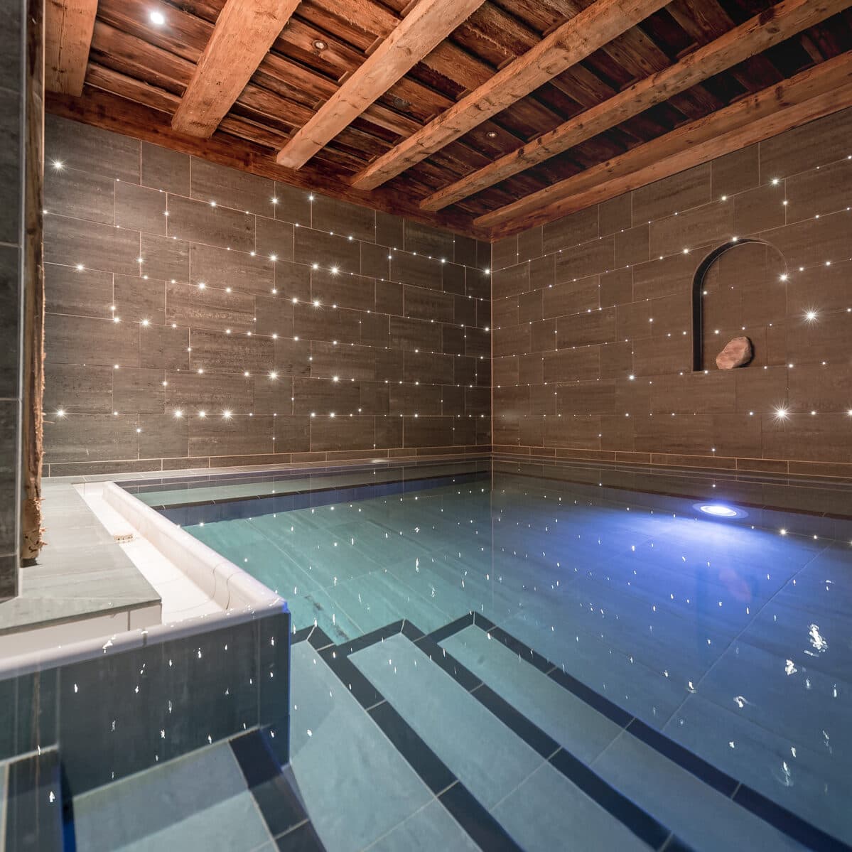 Piscine intérieure avec murs en carreaux de pierre, plafond en bois, niche murale intégrée et lumières scintillantes créant un effet étoilé sur les murs.