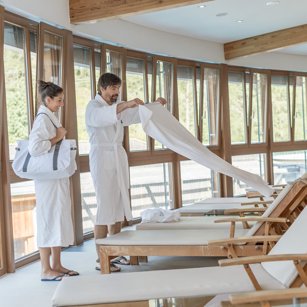 Un homme et une femme en robe blanche se tiennent près de chaises longues à l'intérieur ; l'homme étend une serviette sur une chaise tandis que la femme tient un sac. De grandes fenêtres bordent le mur derrière eux.