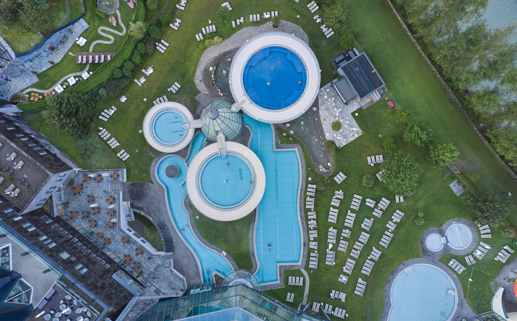Luftaufnahme eines Freibadkomplexes mit mehreren runden und rechteckigen Becken, umliegenden Rasenflächen, Liegestühlen und angrenzenden Gebäuden.