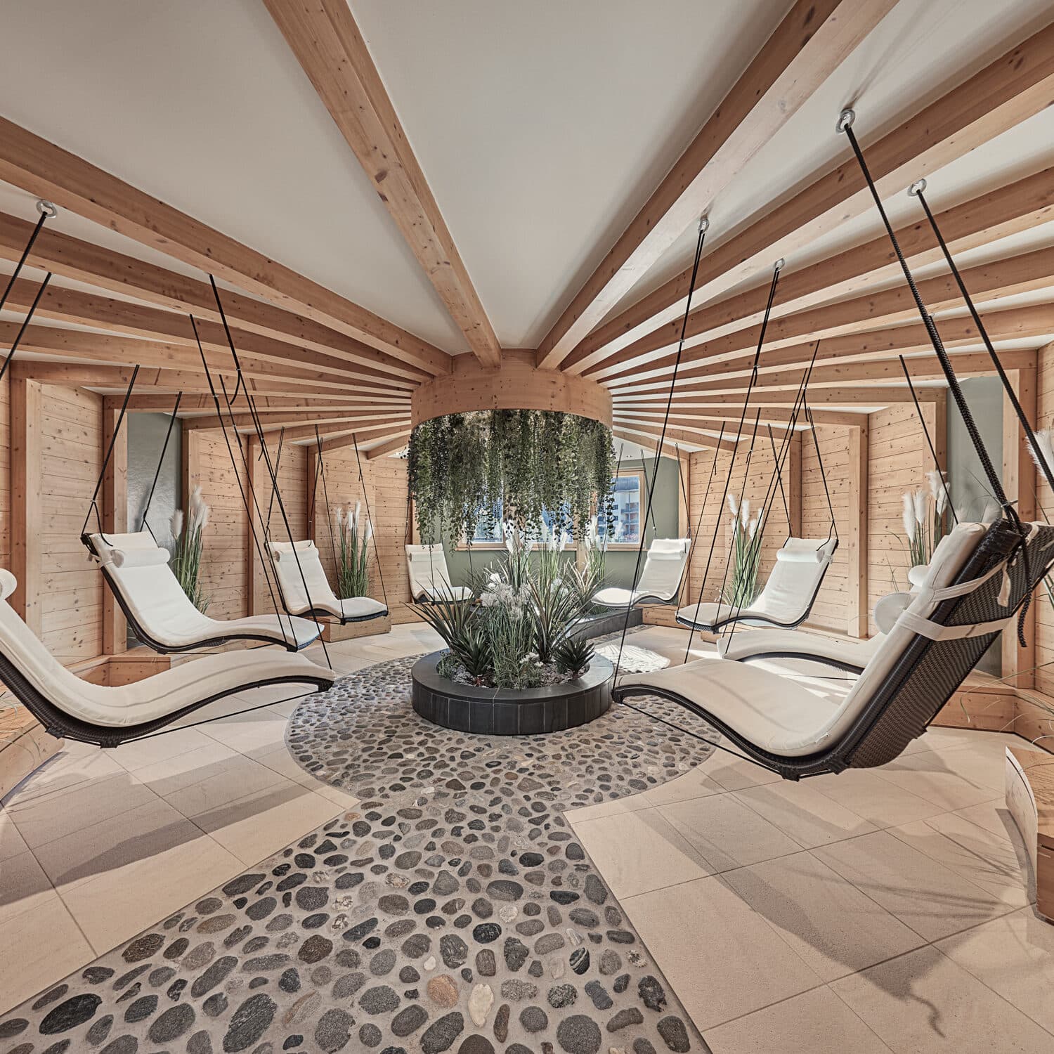 Une pièce circulaire avec des poutres en bois présente des chaises longues blanches suspendues, un sol en galets et une jardinière centrale avec des plantes vertes sous un arrangement de feuillage suspendu.