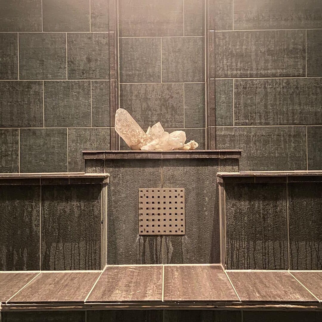 Une grappe de cristaux de quartz clair est posée sur un rebord carrelé dans un hammam aux carreaux sombres et humides, équipé de deux bancs.