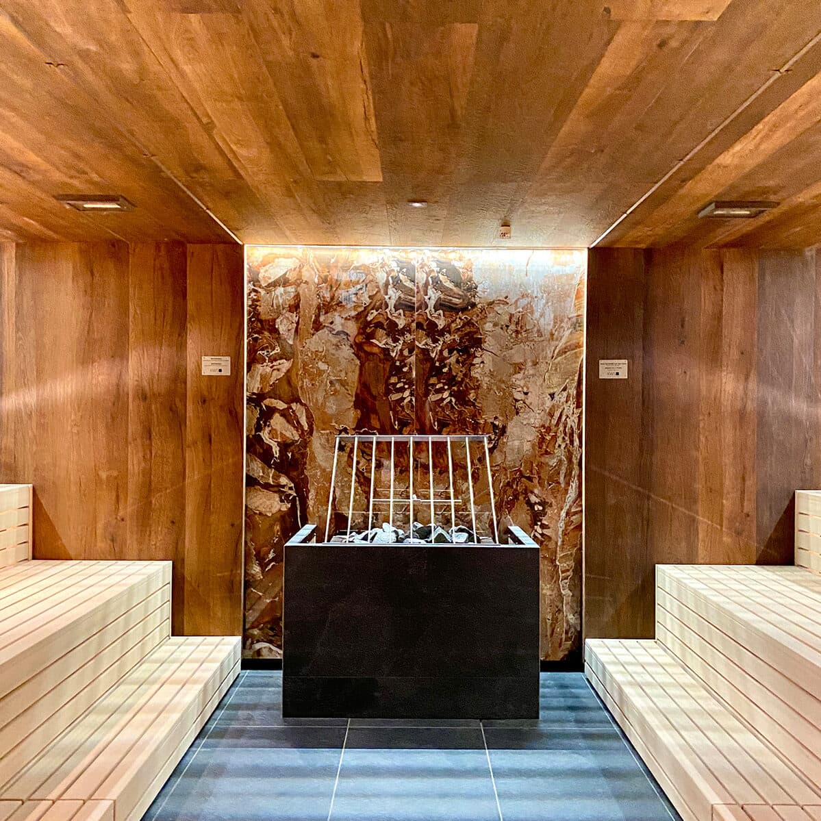 Salle de sauna moderne avec des bancs en bois des deux côtés, un mur en pierre à l'arrière et un poêle central rempli de pierres.