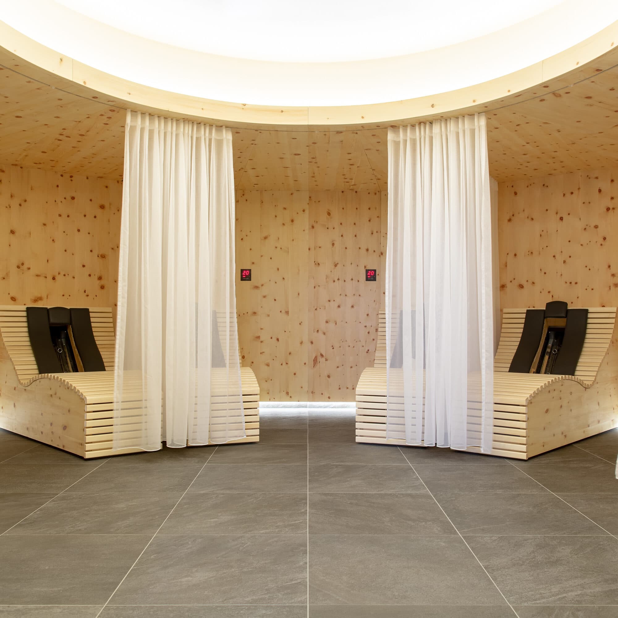 Deux chaises longues en bois séparées par des rideaux blancs dans une salle de détente moderne et circulaire avec un plafond en bois clair et un sol carrelé gris.